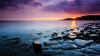 A tranquil seaside scene at sunset, featuring smooth stones along the shore and soft waves under a dramatic, colorful sky