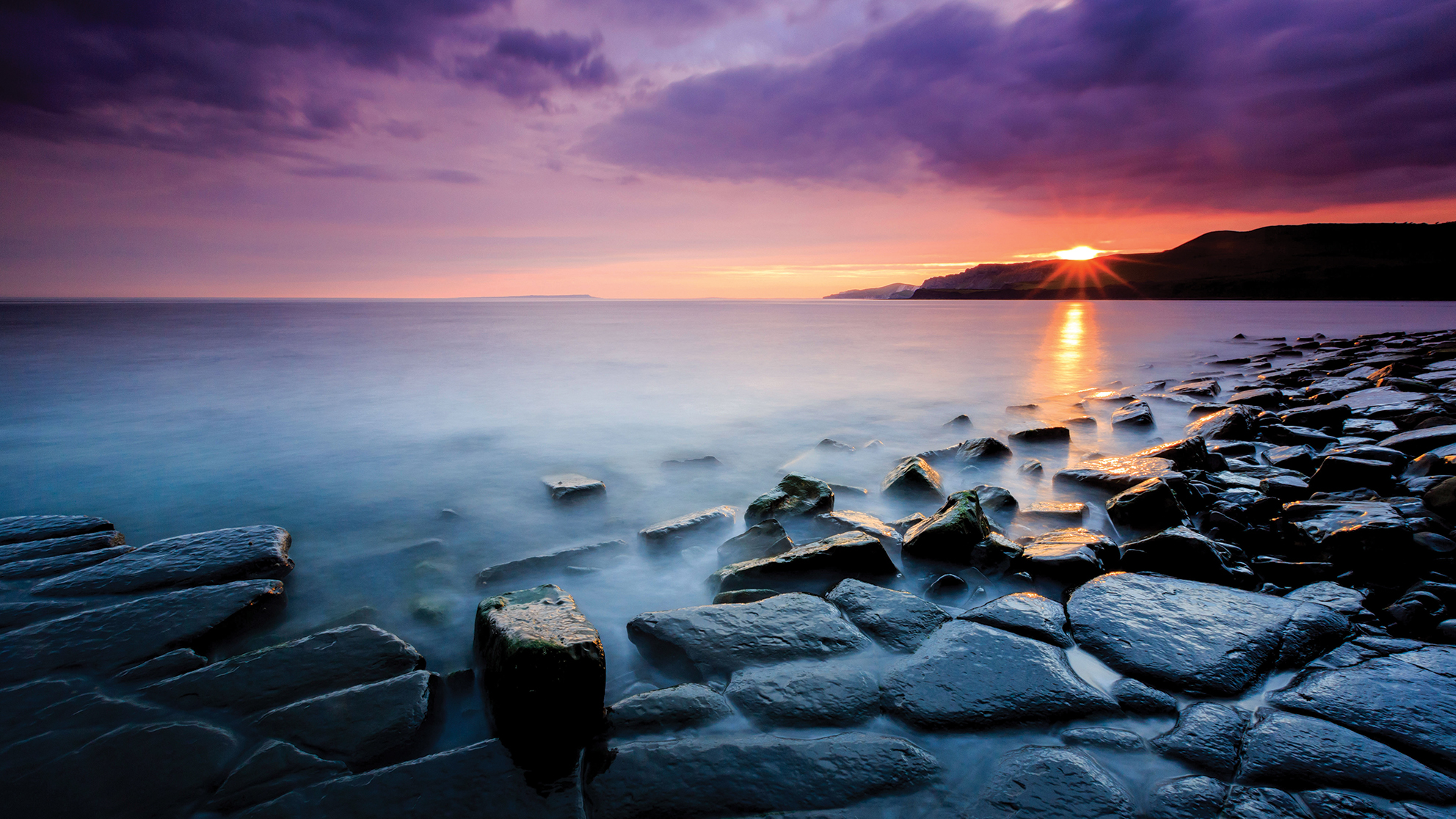A tranquil seaside scene at sunset, featuring smooth stones along the shore and soft waves under a dramatic, colorful sky