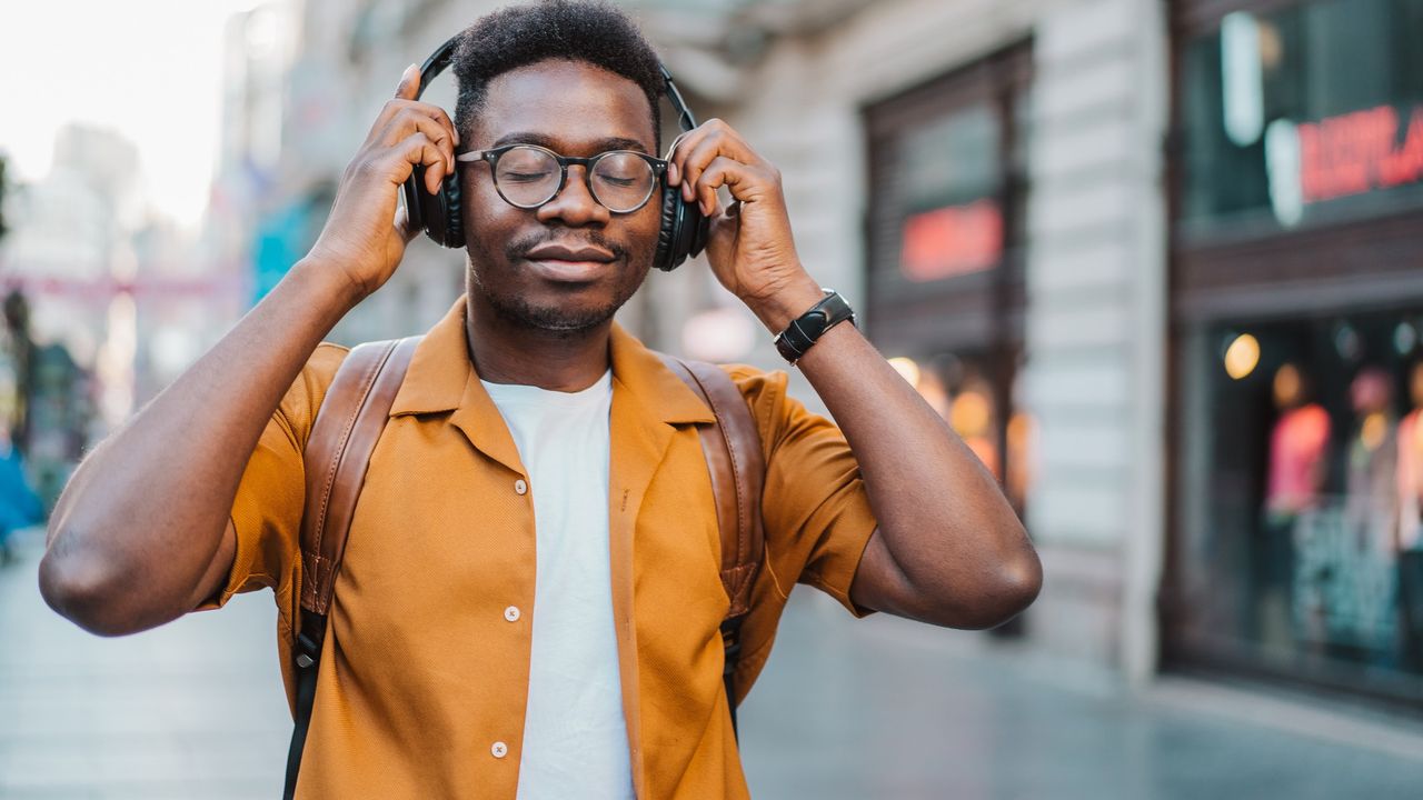 A man wearing round glasses, a mustard shirt and headphones walks through a quiet city street. He has his eyes closed and holds his hands to his headphones. His expression is peaceful.