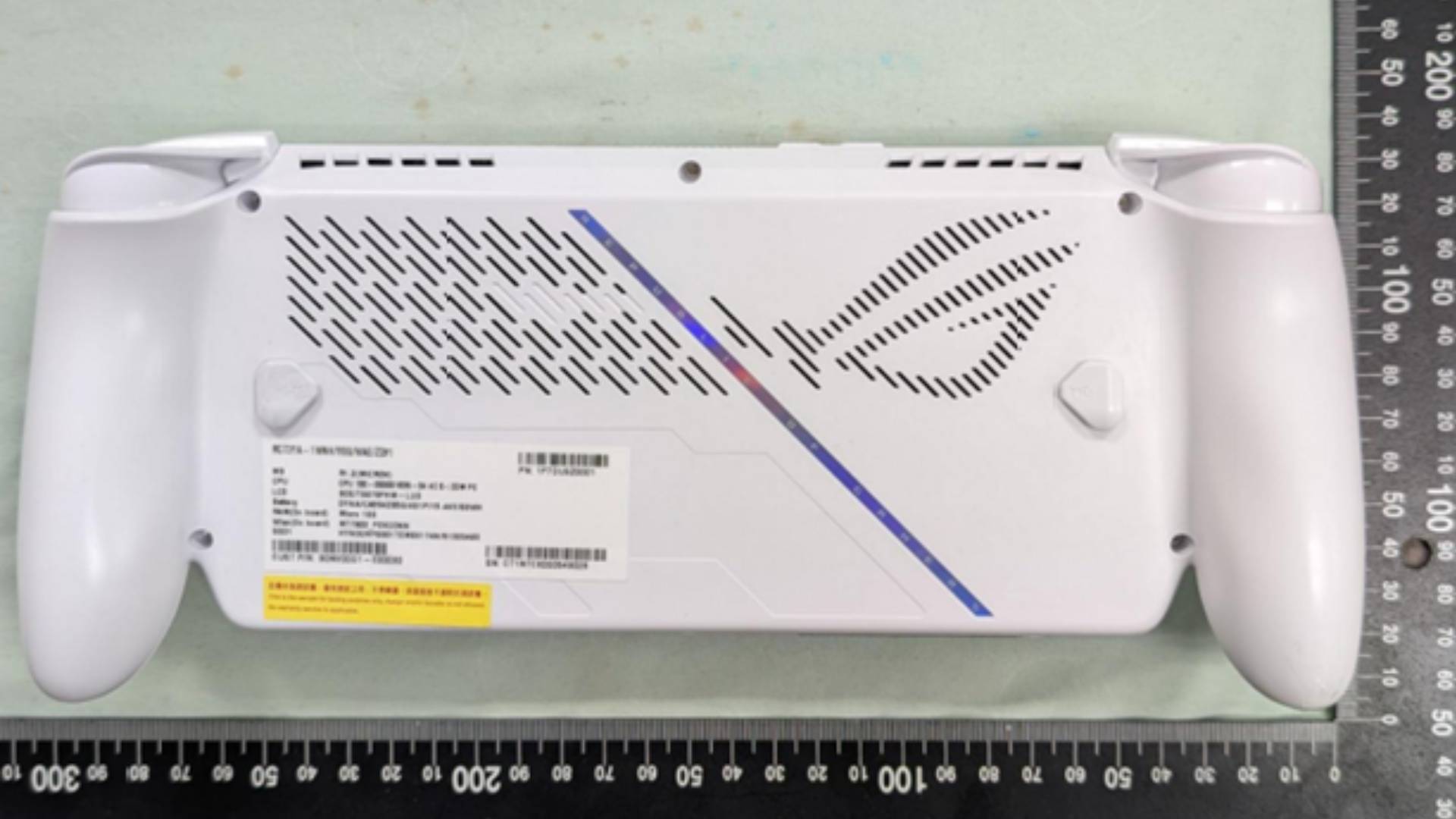 Xbox handheld in white with back facing upwards on light green surface next to ruler.