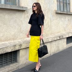 Eliza Huber at Fondazione Prada wearing a black sculpted Carven tunic with a yellow, satin Prada midi skirt, black wedge heels, and a black 1995 Prada bag.