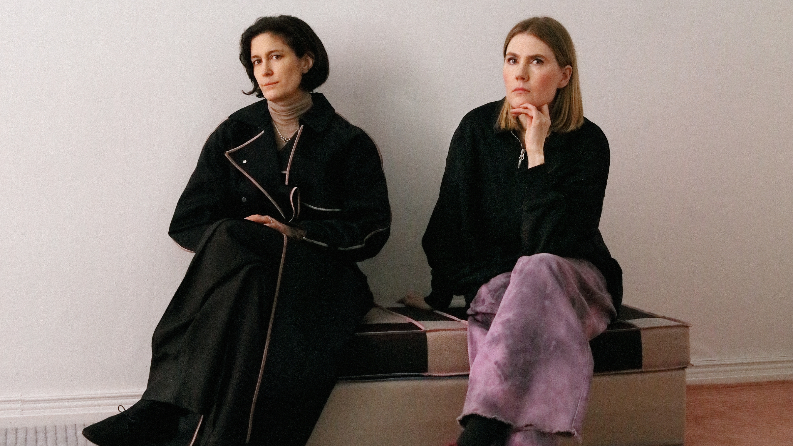 Two young women dressed in oversized black jackets and beige, or pink, clothes sit on an upholstered, checkered bench in textile and chrome in a white room while staring at the camera.