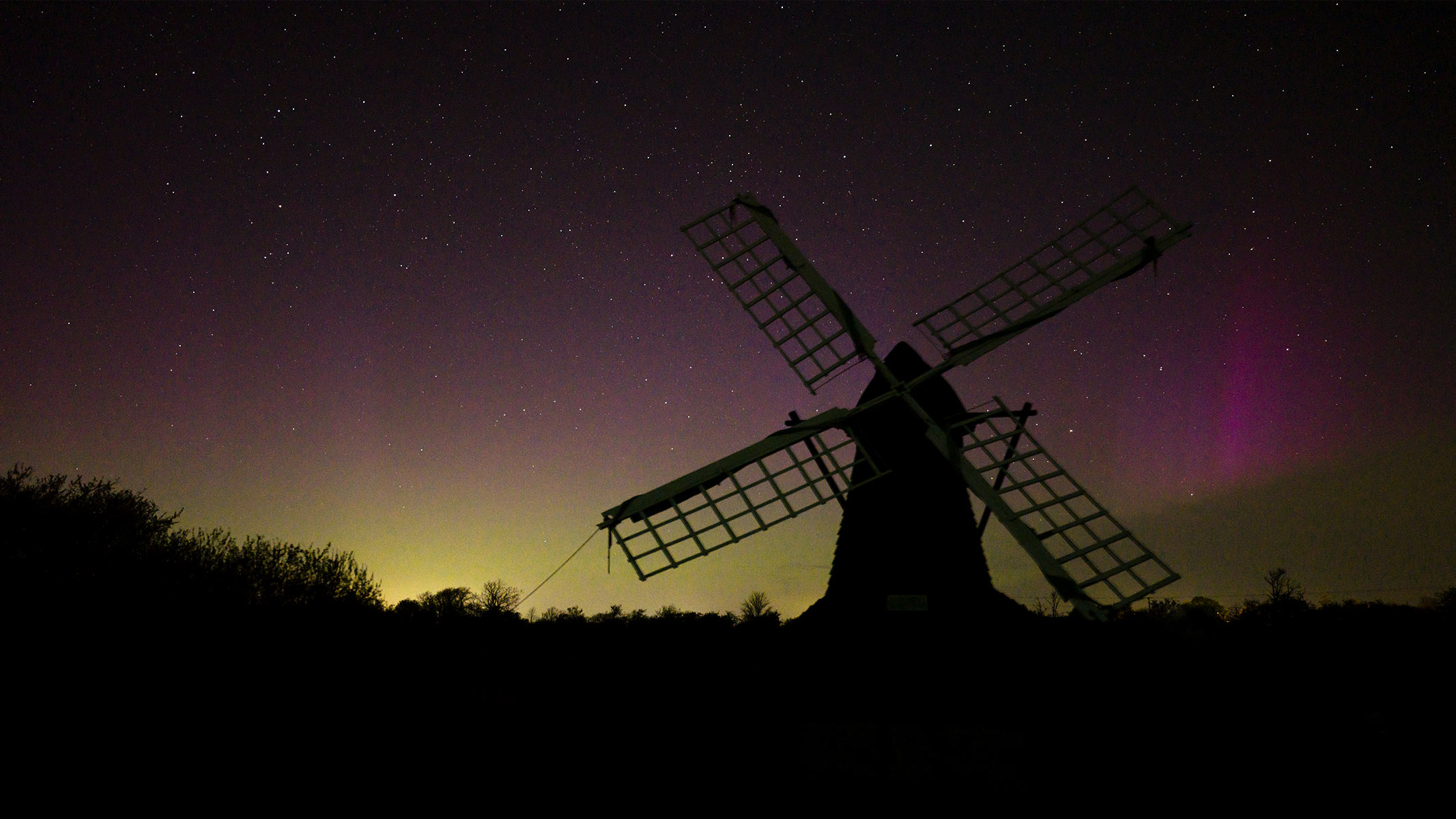 A low-angle shot looking up at a windmill at night with a black, purple and yellow/green color in the background.