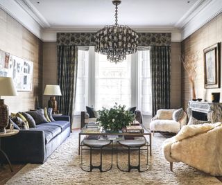 Living room with high ceilings, coving, deep skirting boards and bay window. Dramatic central chandelier, marbled mantelpiece and textured upholstered soft furnishings.