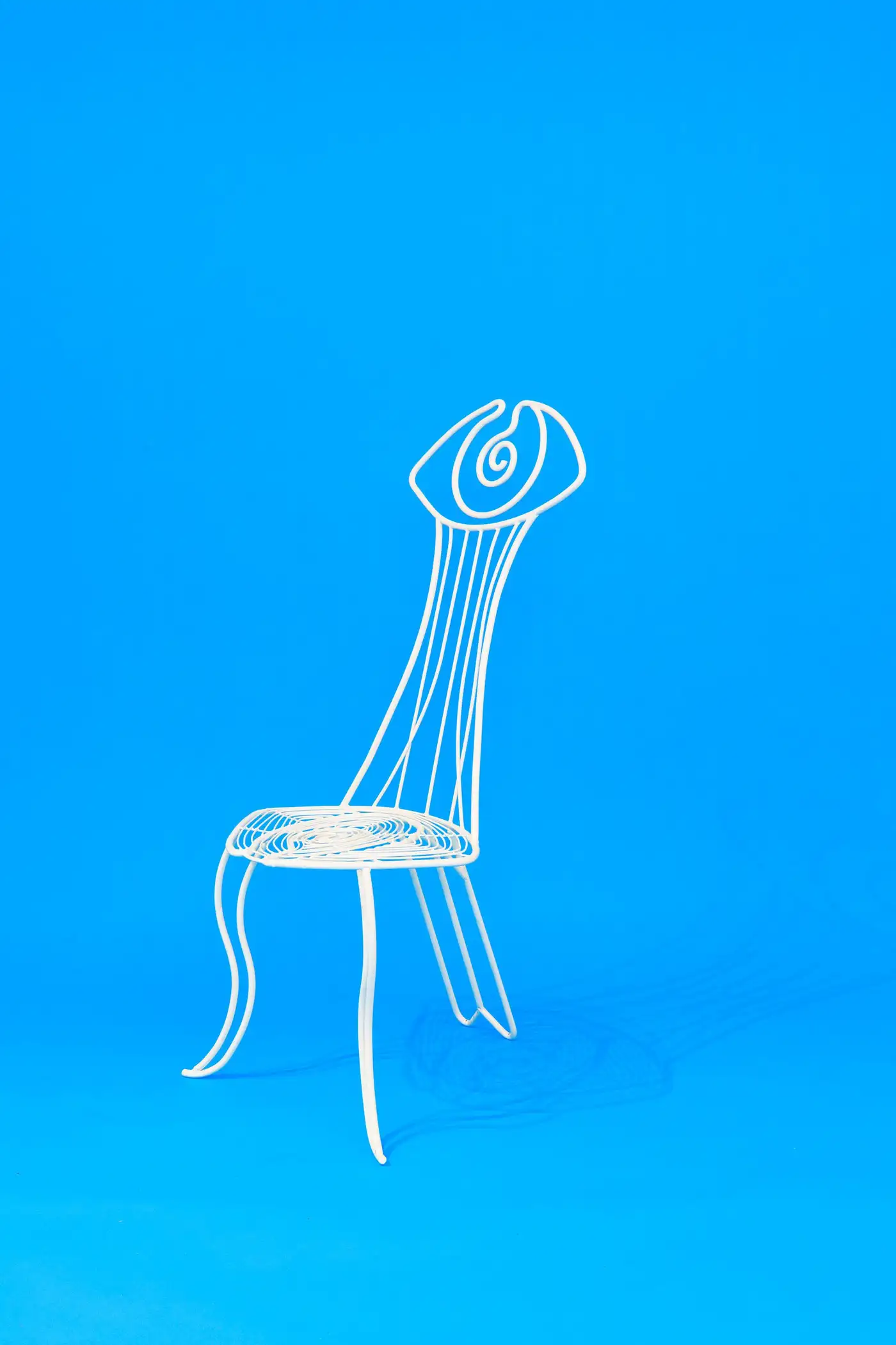 A whimsical chair in painted white wrought iron features an eye motif that makes it creaturesque while sitting against a pale blue background.