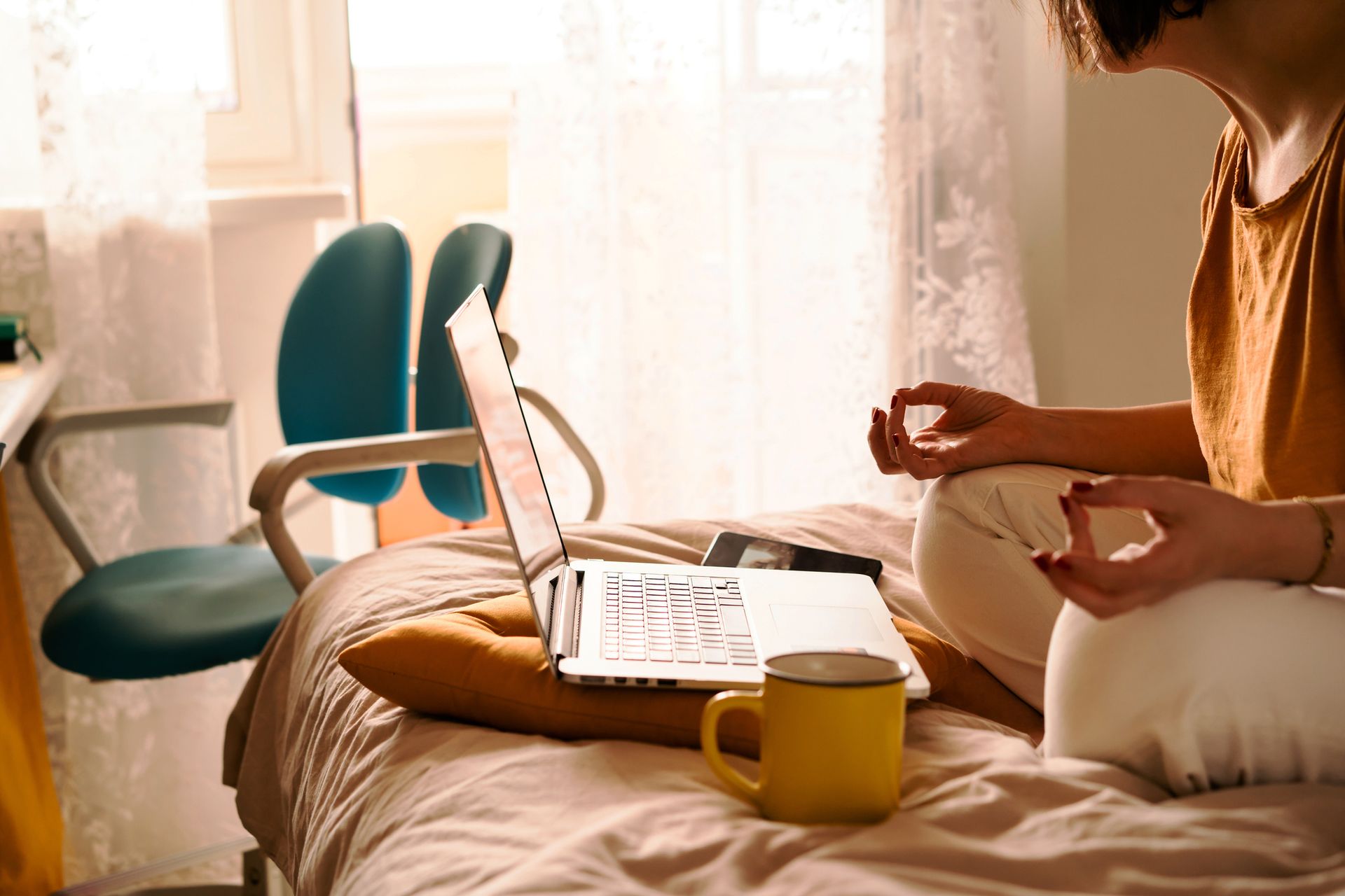 Self care ideas: A middle-aged woman in white jeans and a yellow sweater sitting on the bed in a yoga pose in front of a laptop and a cup of coffee