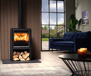 Contemporary freestanding log burner with log storage below, from Charlton & Jenrick, against wood-effect cladding in a living room with a dark blue sofa.