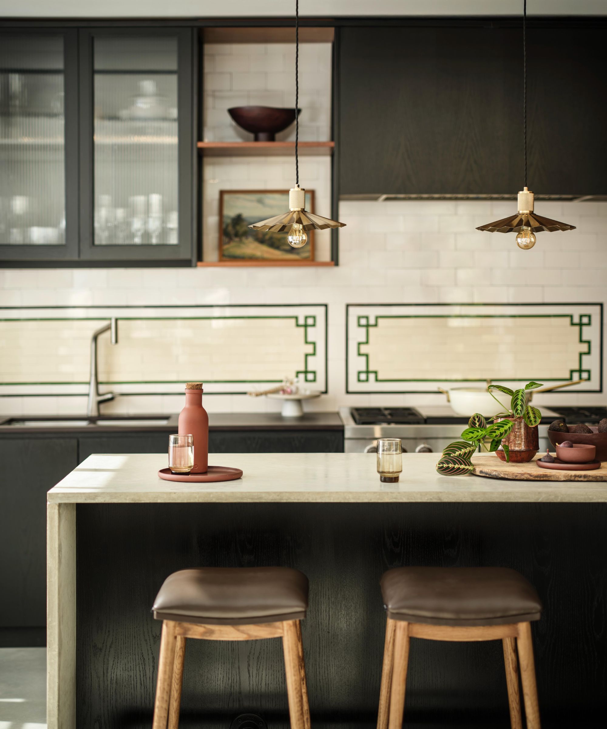 Stone kitchen breakfast bar, with backless stools that have padded leather seats. There is a vintage-looking green geometric design on the far tiled cream wall, with dark green cabinets and open shelving
