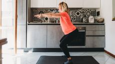 woman performing a squat in her kitchen, sideways to the camera. she's wearing a coral coloured tshirt and black leggings, standing on a black exercise mat in front of some silver kitchen counters.
