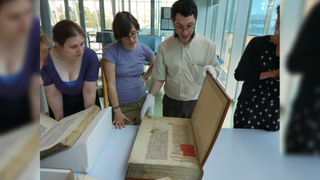 Four people stand in front of a table with a large, old book on top. One wears white gloves and opens the cover.