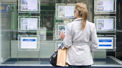 A woman with a shopping bag looks at real estate listings posted on a store window.
