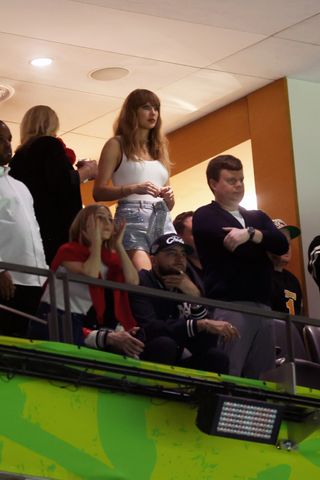 Taylor Swift standing in the audience at the 2025 Super Bowl wearing an alaia bodysuit and bedazzled shorts