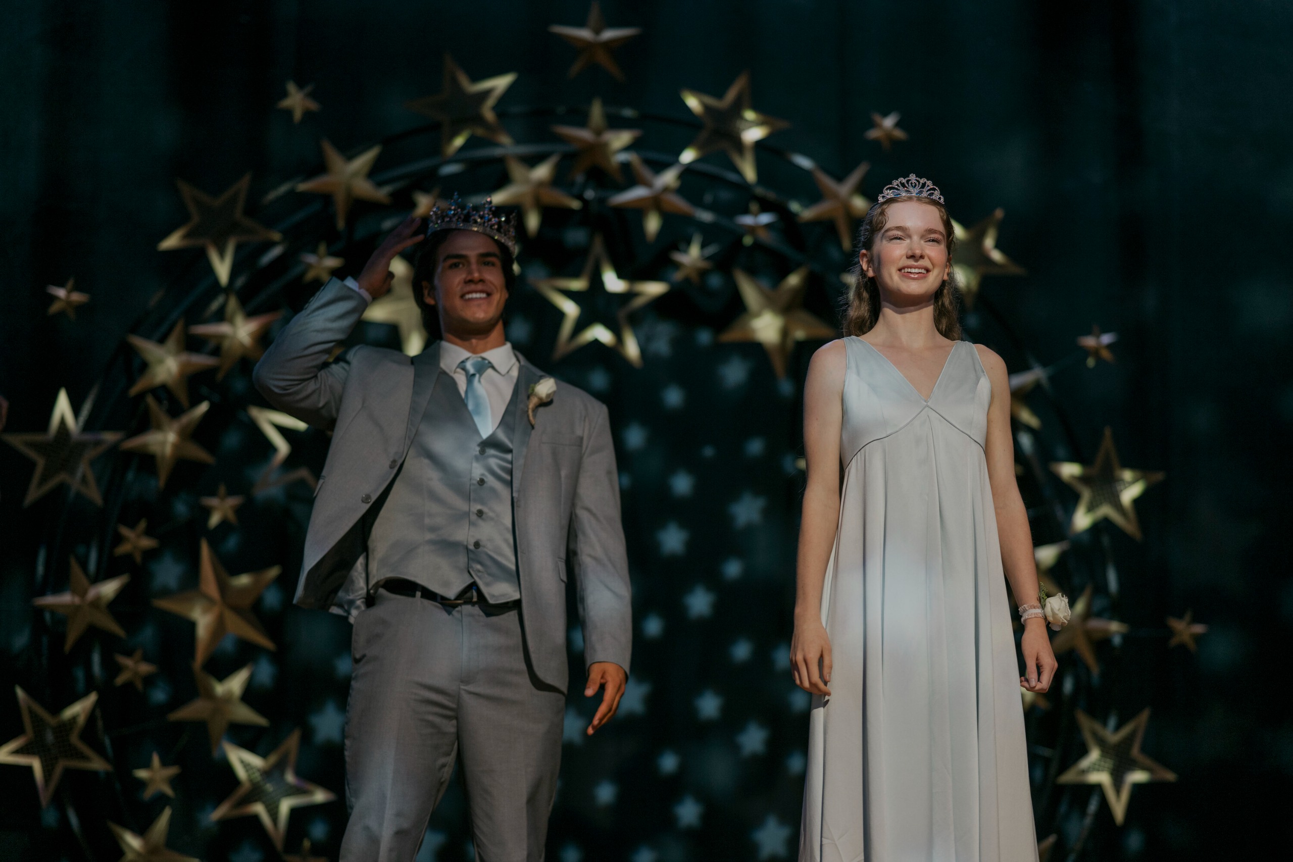 A teenage boy in a gray tux (Joel Oulette as Tommy) and a teen girl in a silver dress and crown (Summer H. Howell as Carrie) both wear crowns as they stand onstage at prom, in Prime Video's 'Carrie.'