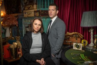 Real-life couple Jessica Ellerby and Nick Hendrix in character as Caitlin Dawson and DS Jamie Winter, standing in an antique emporium surrounded by old suitcases, a champagne bucket, vintage clocks and a classic poker table