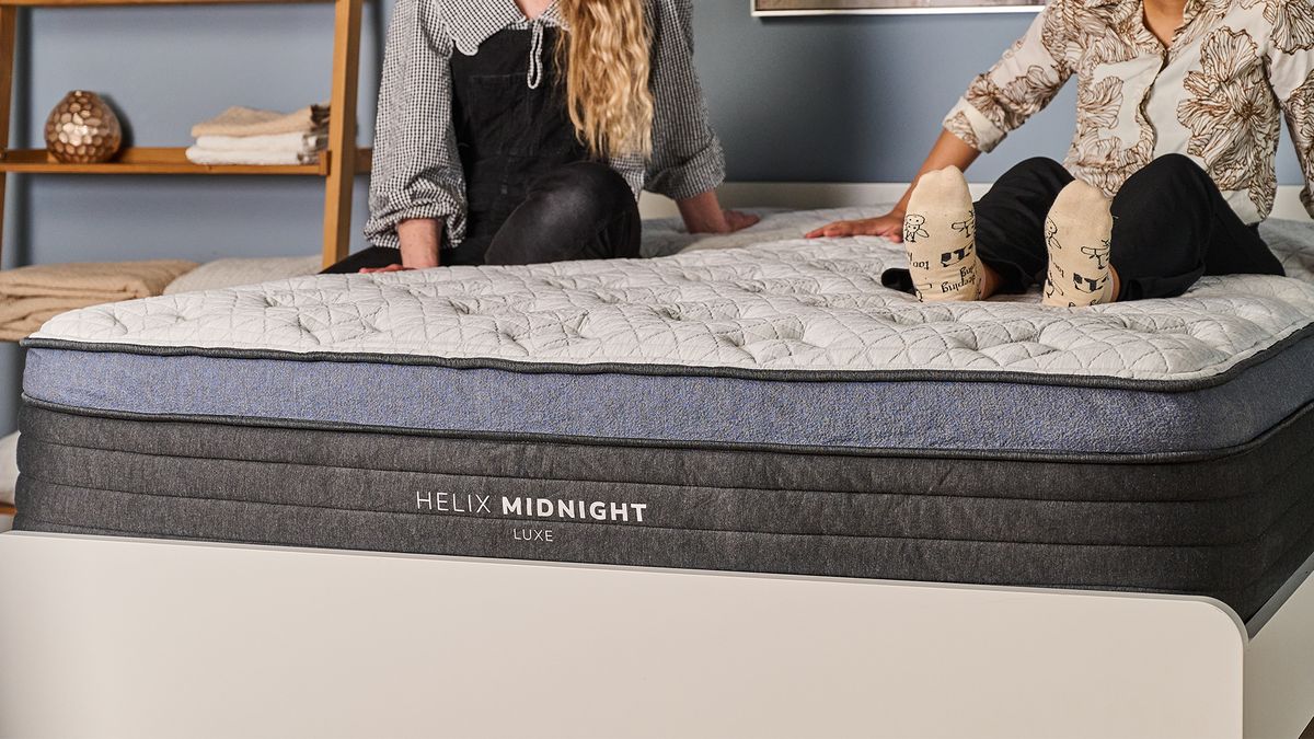 A close up of the foot of the Helix Midnight Luxe Mattress on a bed frame in a bedroom, two mattress testers are sitting on top of the bed