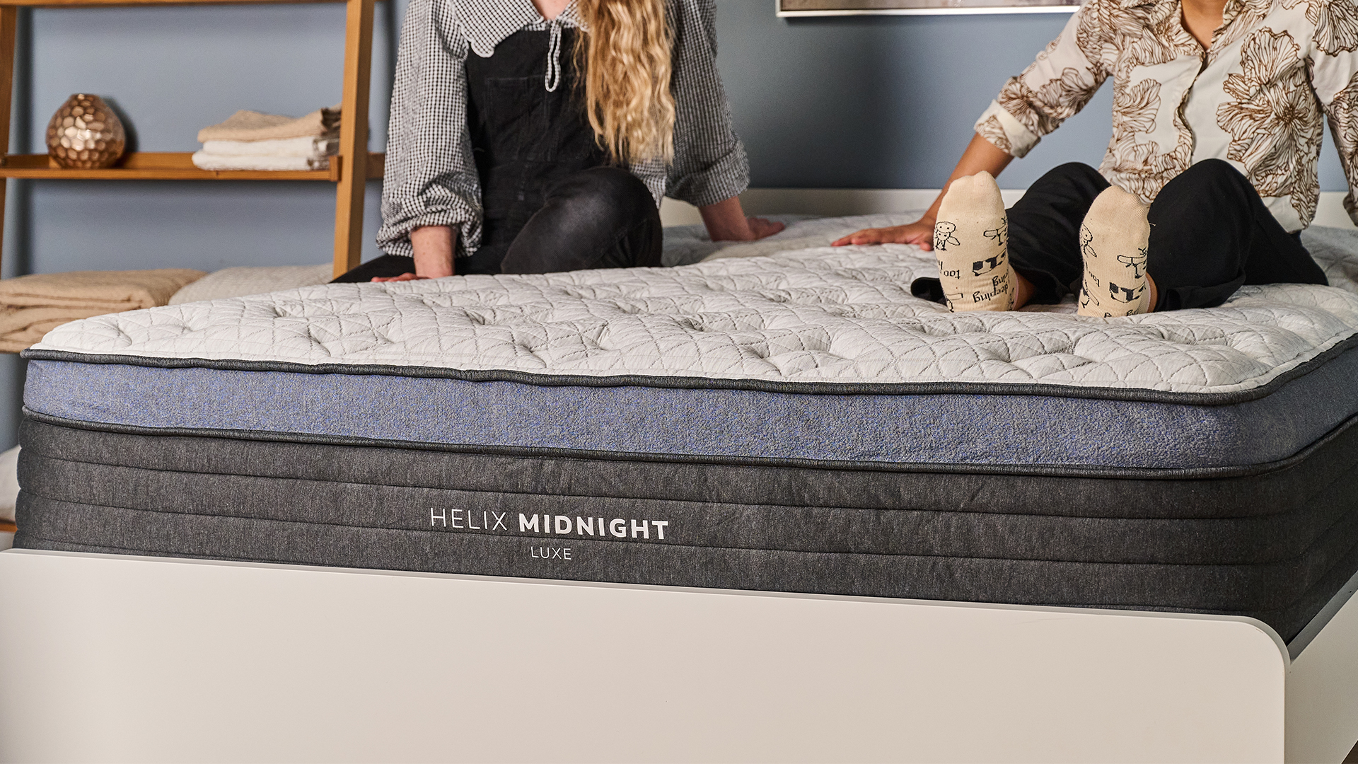 A close up of the foot of the Helix Midnight Luxe Mattress on a bed frame in a bedroom, two mattress testers are sitting on top of the bed