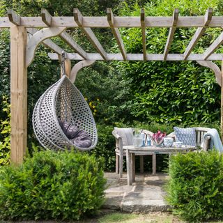 a pergola covered patio in a garden with a hanging egg chair and a small bistro table and chairs