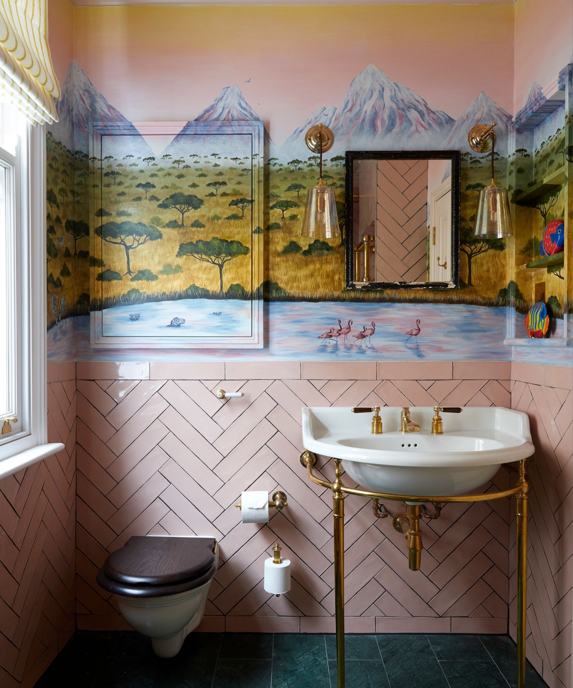A bathroom with a loo and a basin with pink tiles half way up the wall. The upper part of the wall is a colourful painted landscape. On the wall is a rectangular mirror with a black frame and either side two glass wall lights.