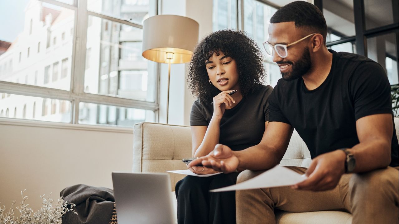 A couple sit next to each other on the sofa and discuss paperwork while looking at a laptop.