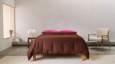 bedroom with window seat, concrete floors, beige rug, beige and silver armchair, side table with paper lantern, stack of magazines, bed made with brown and fuschia pink bedding