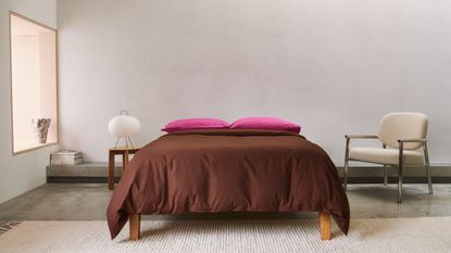 bedroom with window seat, concrete floors, beige rug, beige and silver armchair, side table with paper lantern, stack of magazines, bed made with brown and fuschia pink bedding