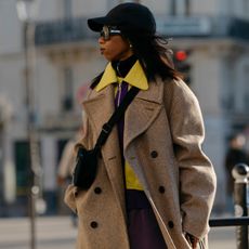 Attendee at paris fashion week menswear fall winter 2023 wearing camel coat, yellow shirt, crossbody bag, black hat, and sunglasses 