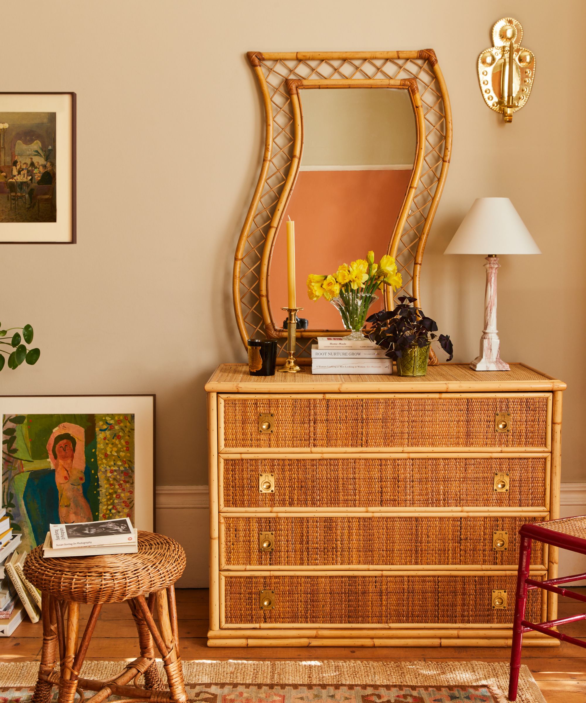 A textured bamboo dresser topped with a unique, wavy-framed rattan mirror, yellow daffodils in a glass vase, and a pink marble lamp against a beige wall.
