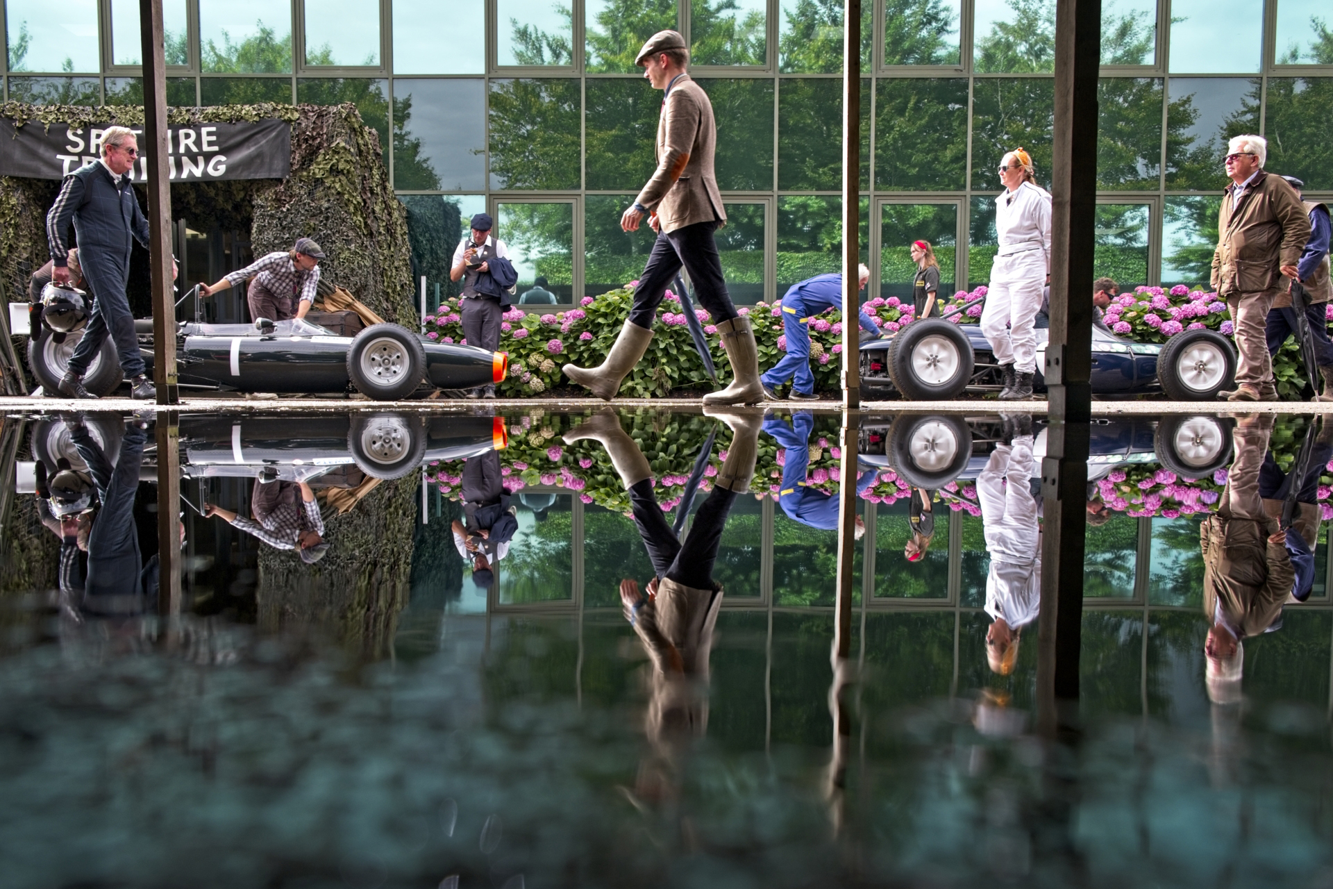 A sleek vintage race car is being prepared in a vibrant garden setting, reflecting beautifully in the glossy surface beneath