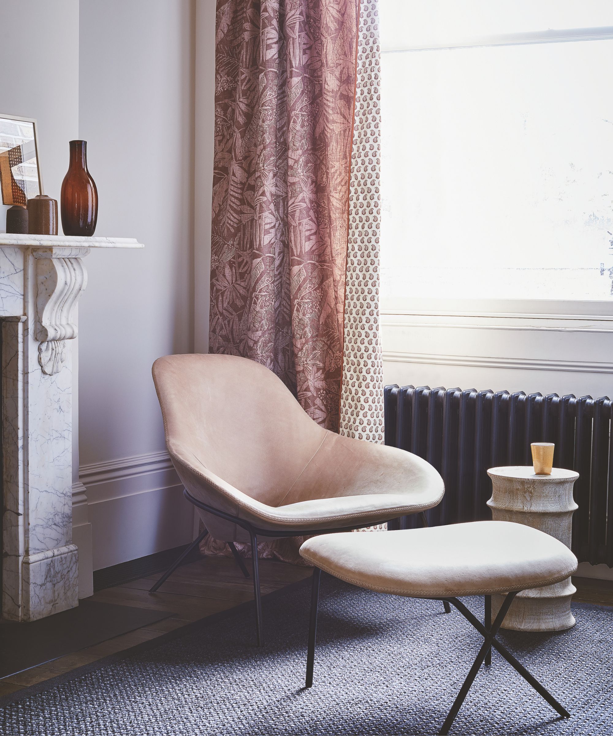 Chair and footstool by a window with a black radiator, pink curtain and marble fireplace