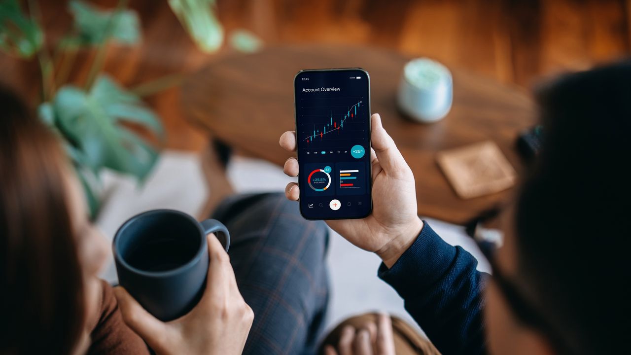 A man&#039;s hand holds up a smartphone, which shows a stock trading graph.