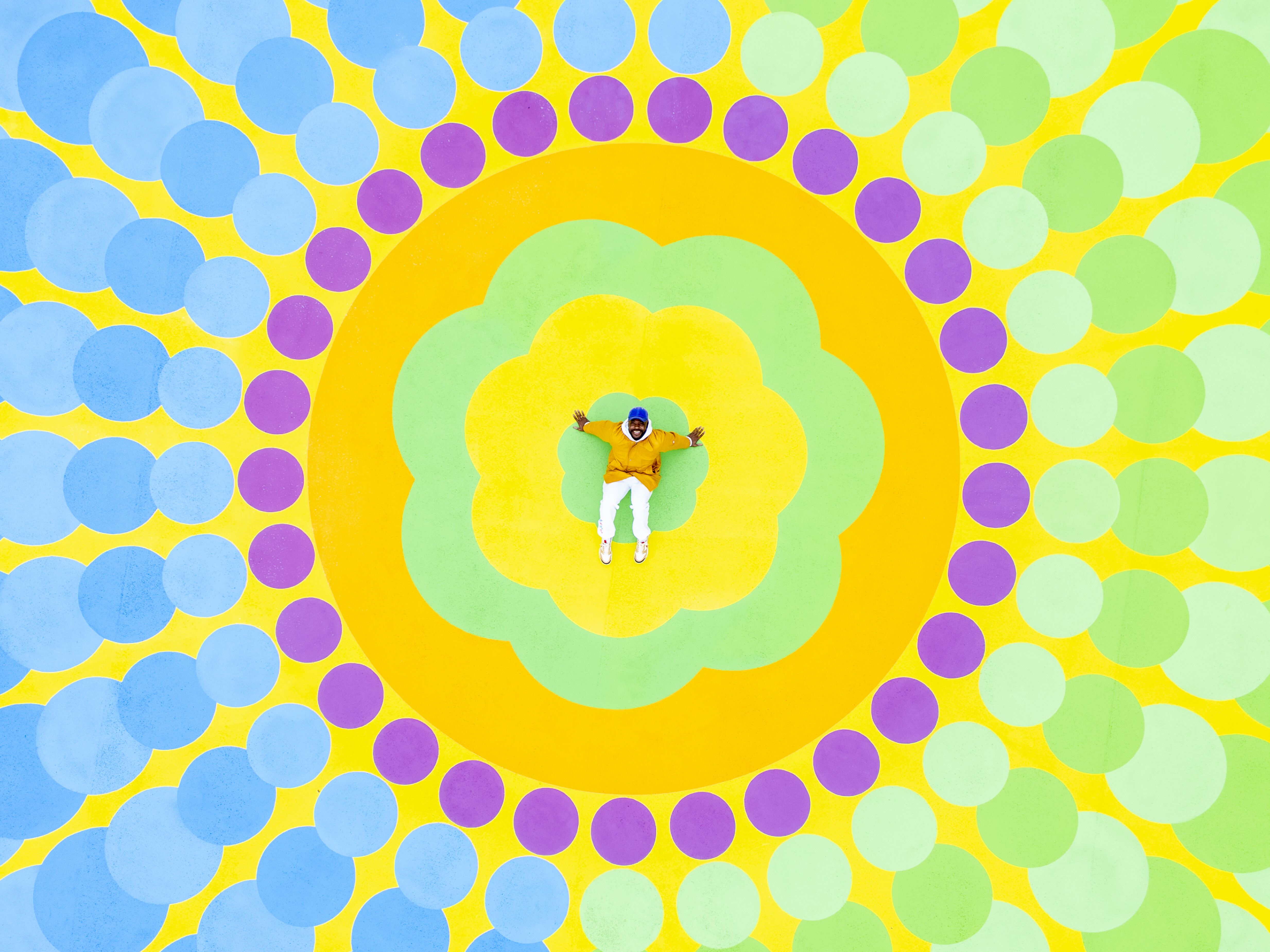 A man dressed in a yellow jacket and white trousers with a blue cap sits at the center of a flower-shaped, large-scale mural in bold colors.