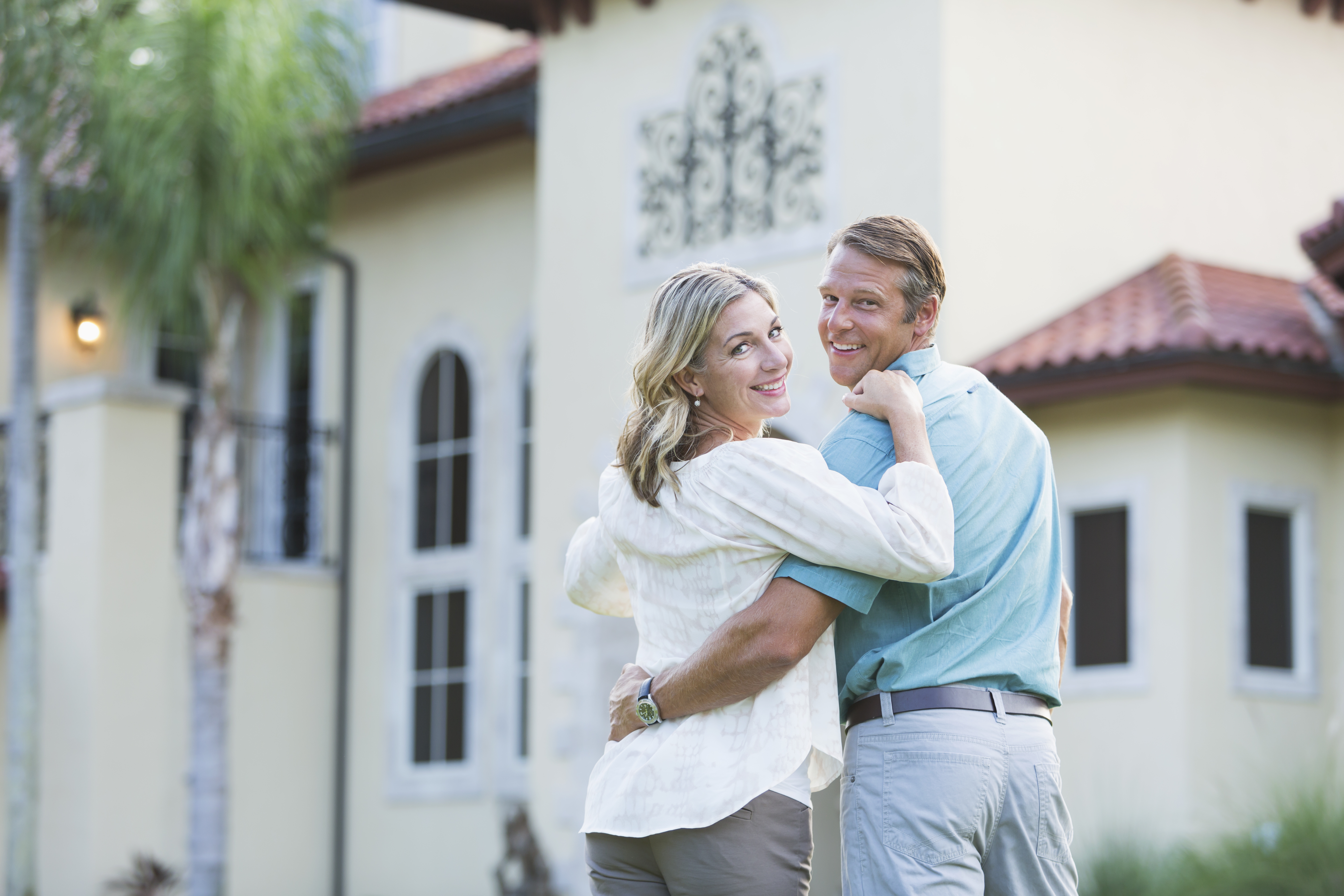 An affluent mature couple standing in front of a home.  They are standing side by side with their arms around each other, looking over their shoulders at the camera.