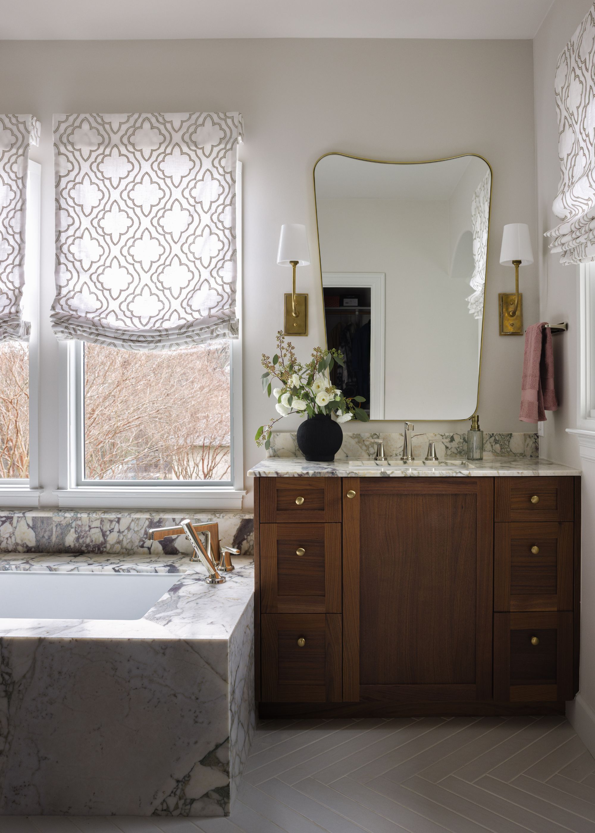 An elegant bathroom featuring a wood vanity with gold hardware and a white marble countertop. A large, irregularly shaped gold-framed mirror is flanked by two brass sconces. To the left, a built-in soaking tub is encased in the same heavily veined marble, set beneath windows with patterned Roman shades.