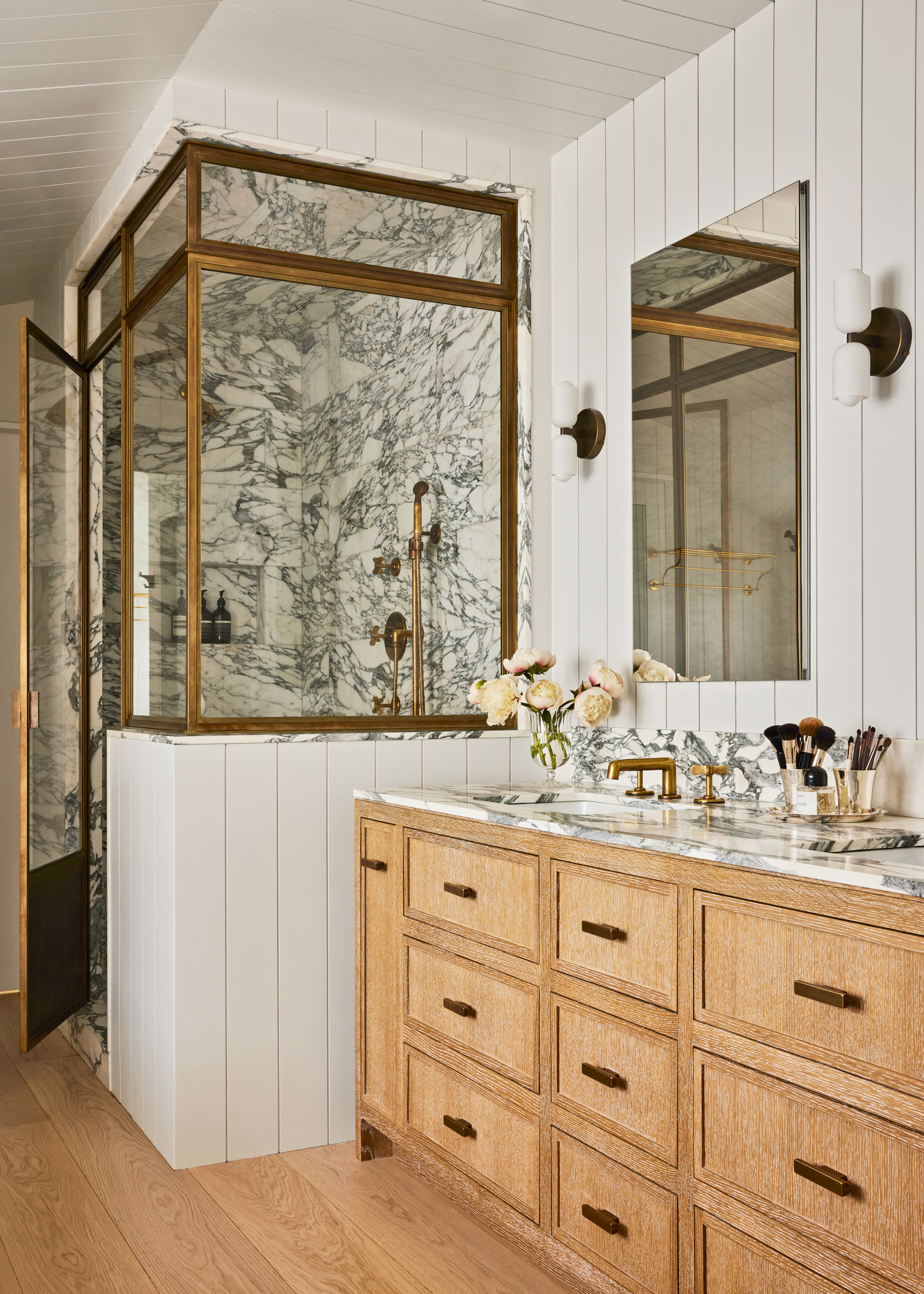 Bathroom with white panelled wall, grey and white veined marble shower and sink countertop, wood vanity and wall lights