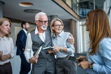 Senior and younger colleagues smiling and embracing in a modern office, sharing mentorship, teamwork and collaboration while discussing projects and strategy in a friendly professional setting