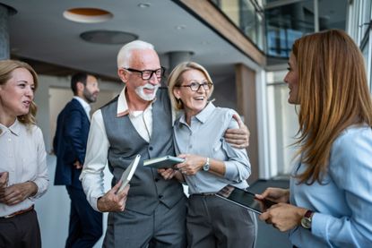 Senior and younger colleagues smiling and embracing in a modern office, sharing mentorship, teamwork and collaboration while discussing projects and strategy in a friendly professional setting