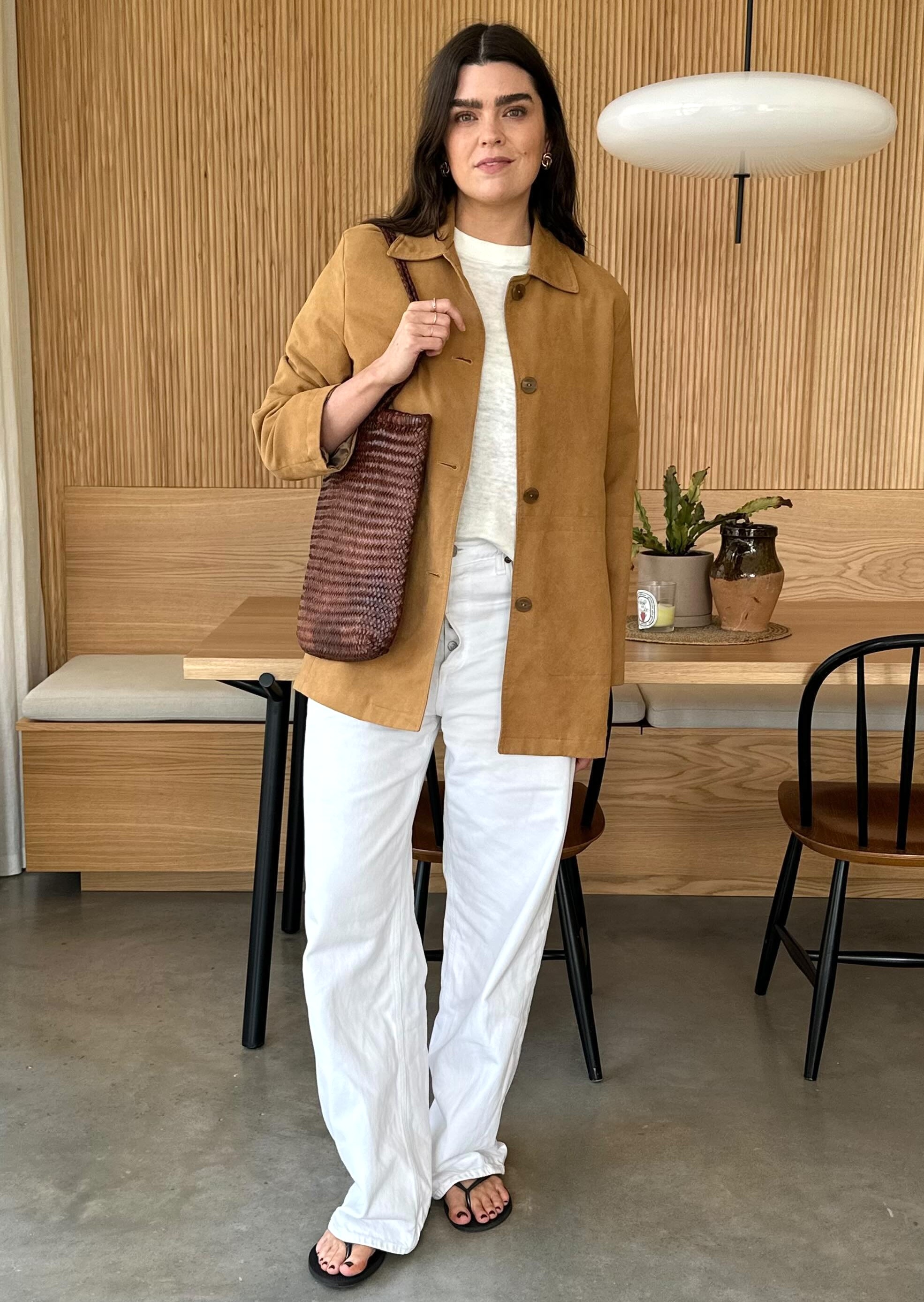 British style influencer Anna Newton poses in her London dining room wearing a tan suede shirt jacket, brown leather woven tote bag, a linen sheer white T-shirt, baggy white jeans, and black flip-flop sandals