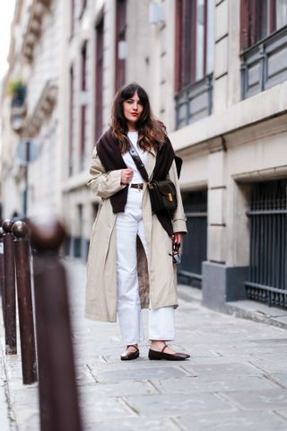 Sarah Benziane wears a white t-shirt, a brown pullover over the shoulders, a beige trench coat from Mango , a black leather belt, white denim pants / jeans from Reiko, a brown leather bag from Leo & Violette, brown leather ballet / ballerina flat shoes, during a street style fashion photo session, on April 03, 2024 in Paris, France.