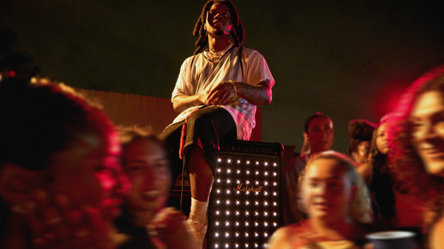 A musician with long locs sits atop a glowing Marshall speaker stack, performing for a crowd at an outdoor nighttime event.