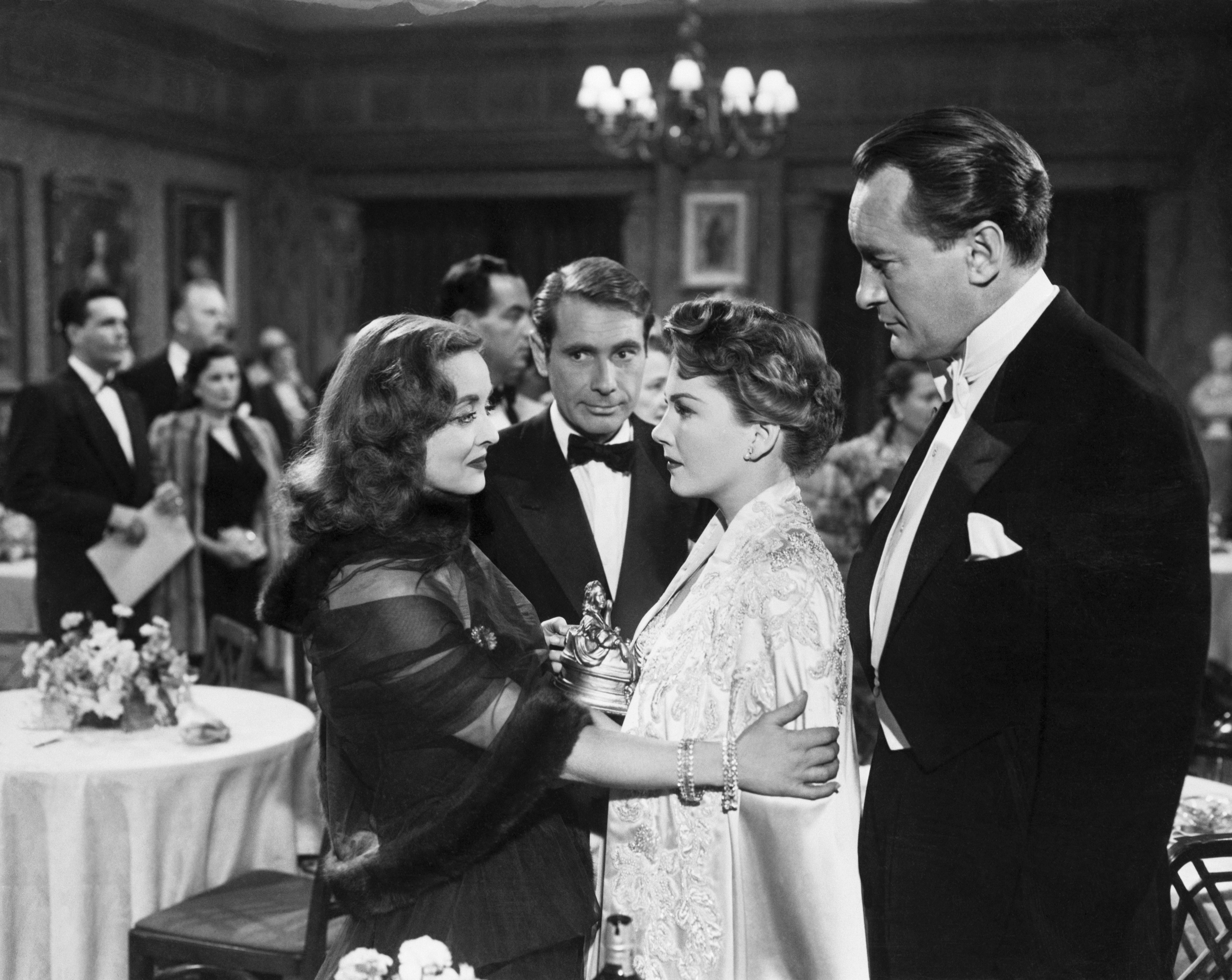 A woman (Bette Midler as Margo Channing) loosely hugs another woman (Ann Baxter as Eve) as two men (Gary Merrill and George Sanders) stand with them, in a still from the 1950 film 'All About Eve.'