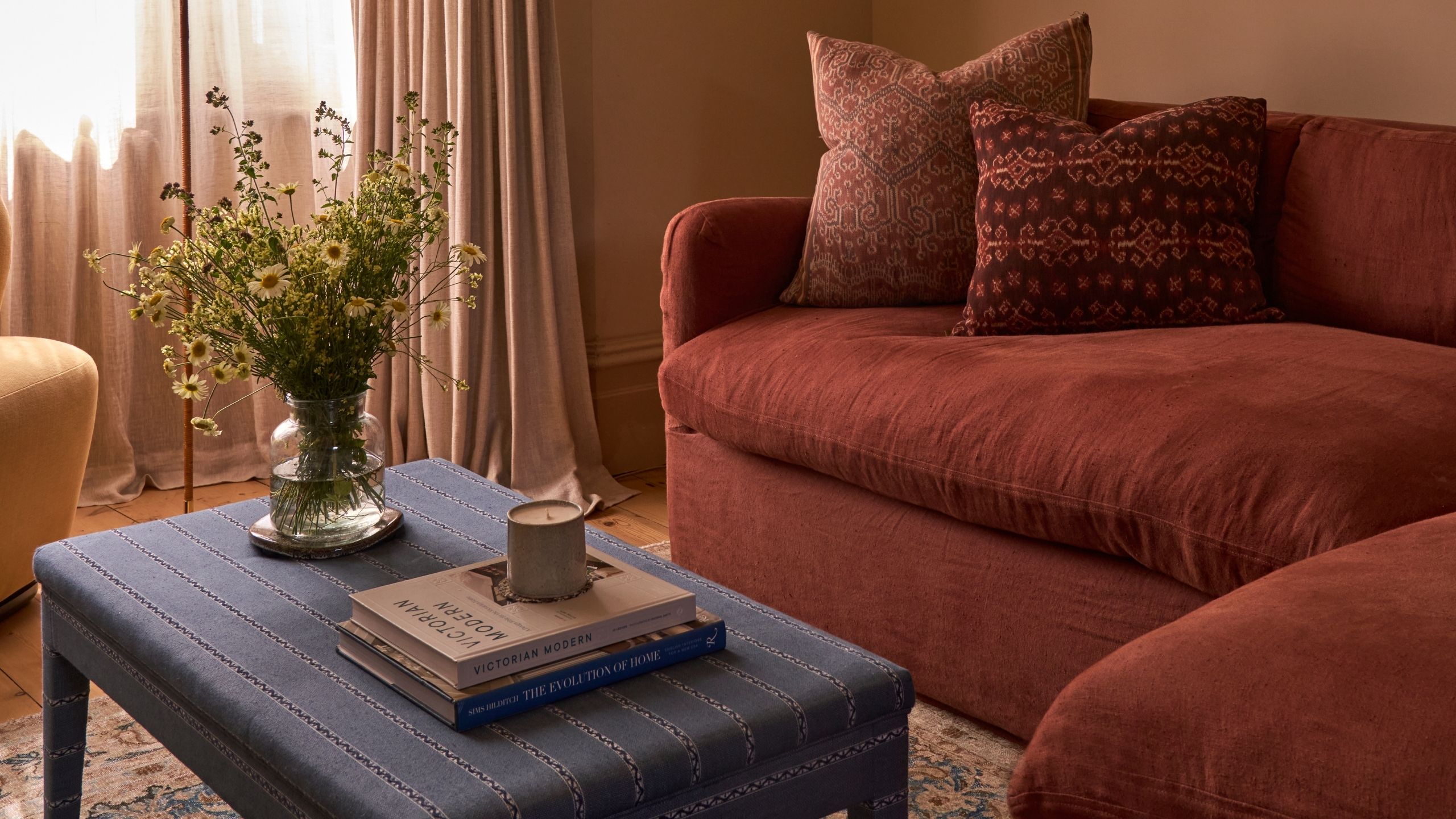 Image of a warm, beige living room with a red sofa, a blue patterned sofa, and a blue upholstered ottoman. 