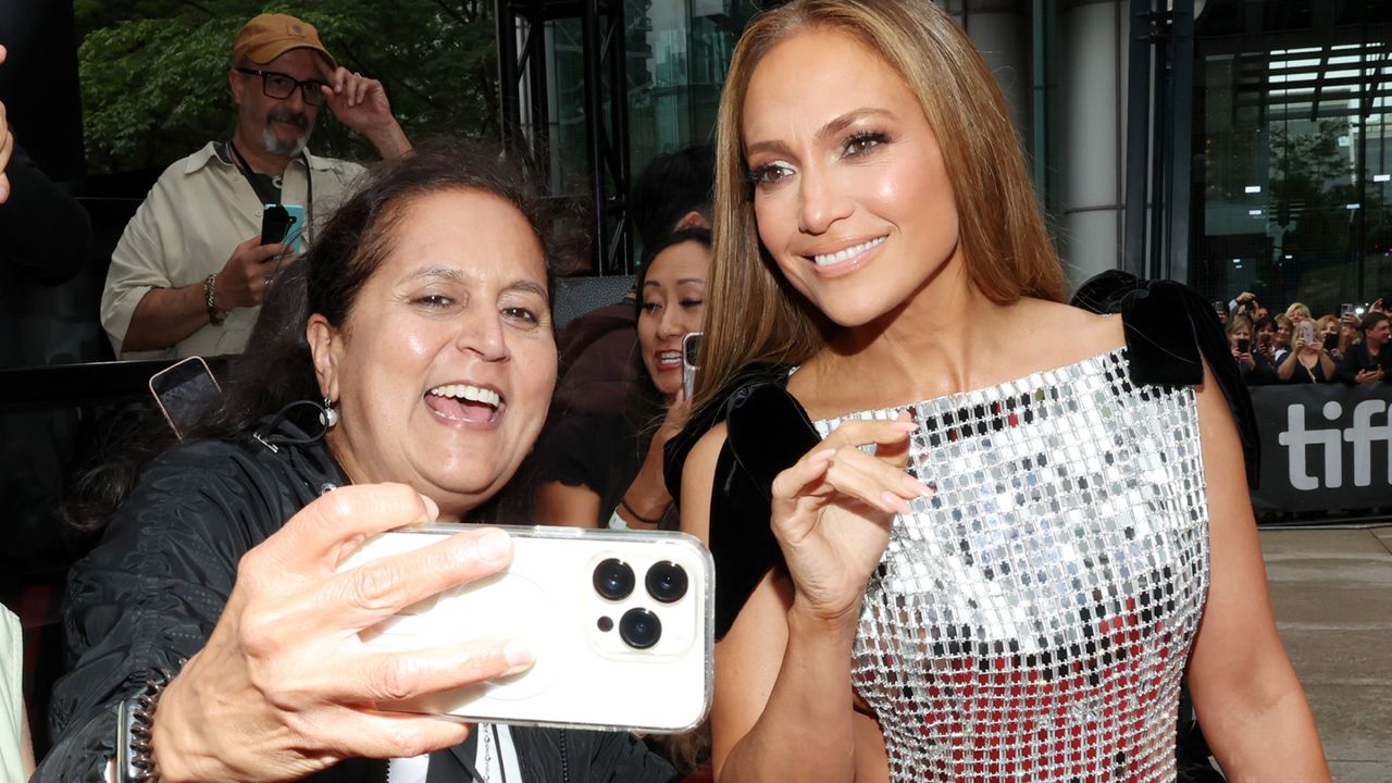 Jennifer Lopez wearing a silver dress and smiling for a selfie with a fan