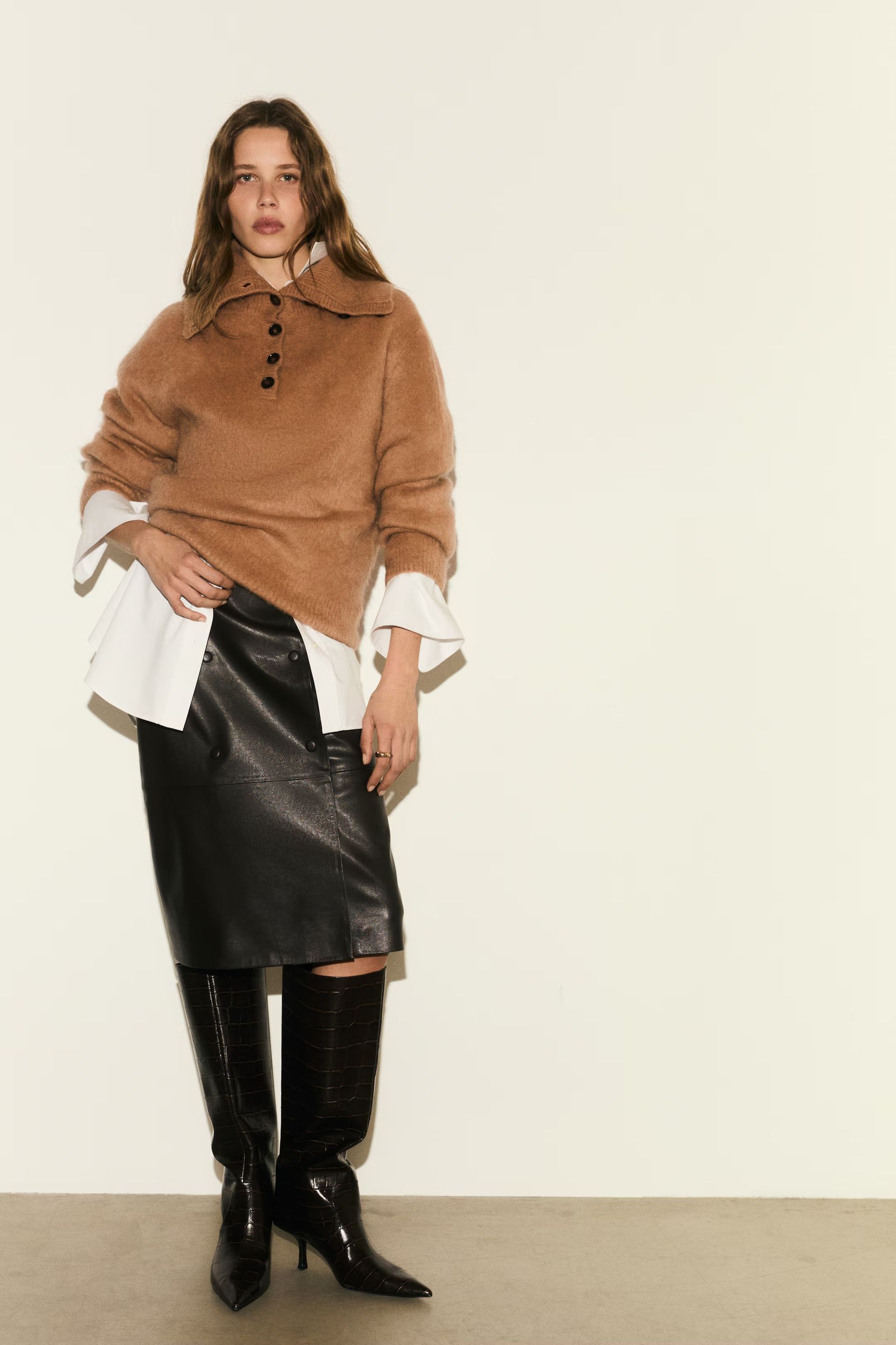 Woman wears beige funnel neck cardigan, leather skirt and knee high boots