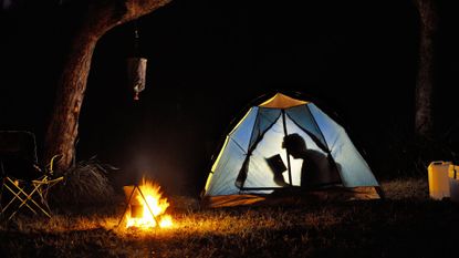 A person sitting in their tent reading a book with a torch