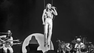 Genesis singer Peter Gabriel is suspended in midair during a concert in New York City, 6 May 1974. Guitarist-bassist Mike Rutherford is seated at left, drummer Phil Collins is behind the kit at right.