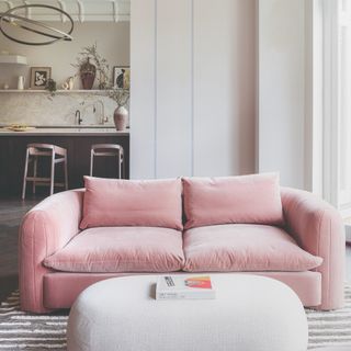 A modern living room with a pink two-seater sofa and a curved coffee table with a kitchen in the background