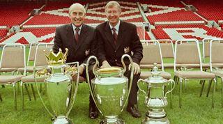 MANCHESTER, ENGLAND - AUGUST 1999: (L-R) Director Maurice Watkins and Sir Alex Ferguson of Manchester United pose with the Premier League Trophy, the UEFA Champions League trophy and FA Cup trophy during the Manchester United squad photocall 1999/2000 session at Old Trafford in Manchester, England. (Photo by John Peters/Manchester United via Getty Images)