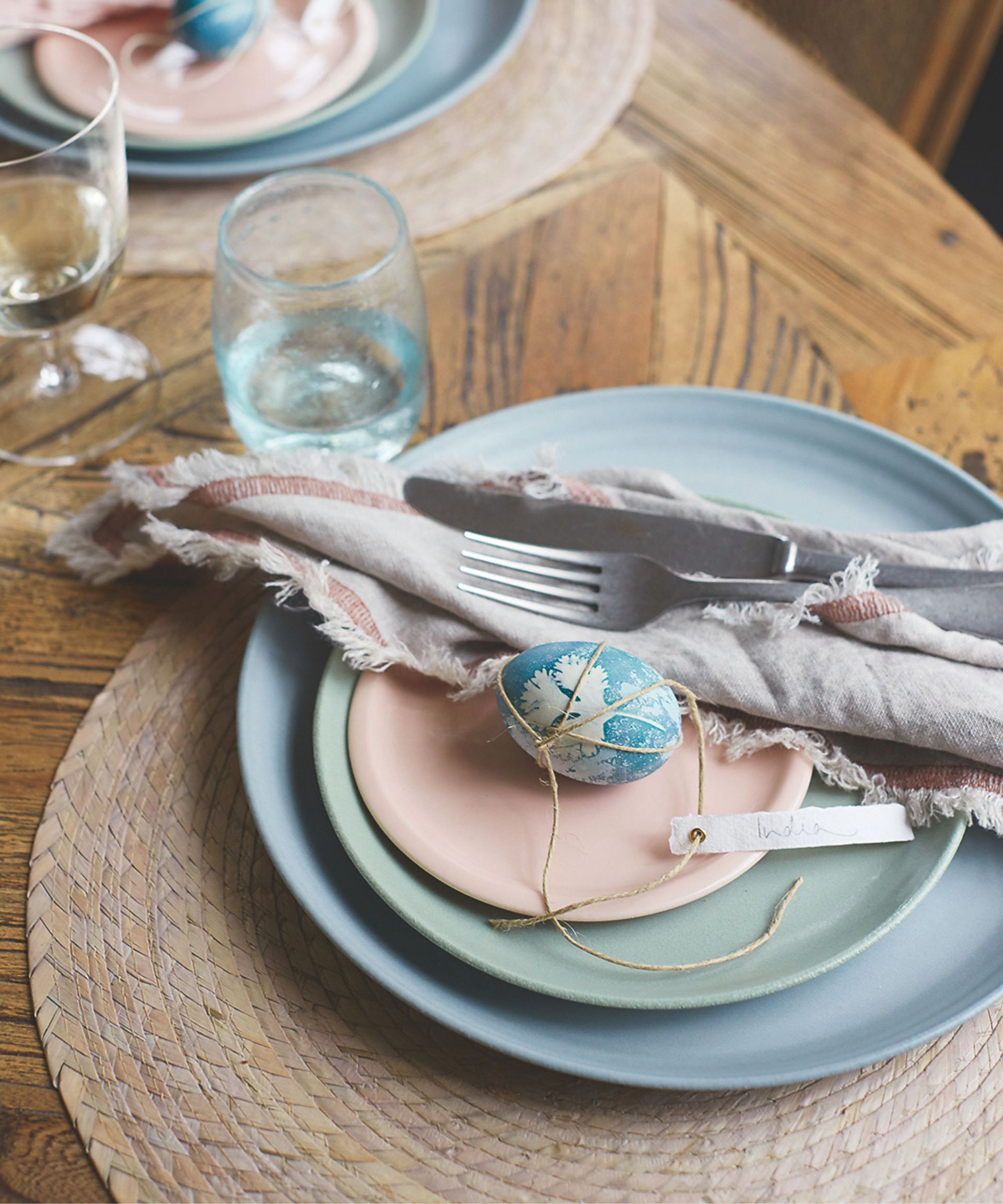 Blue and white decorated egg with a paper tag tied on with brown string, on a stack of pastel coloured places, next to a neutral napkin and set of cutlery
