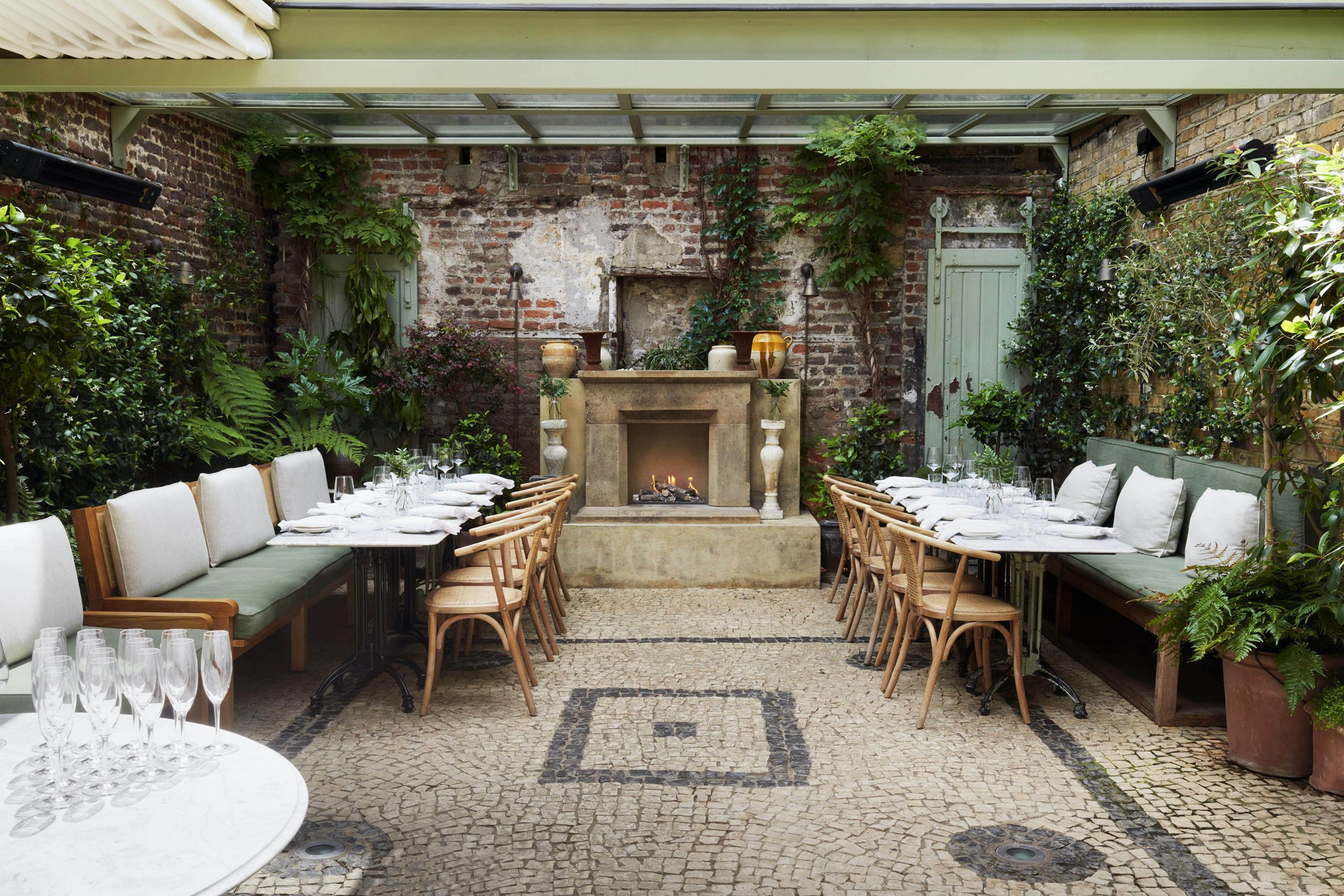 A wide shot of an open terrace with cobblestoned floors and a pale stone fireplace centrepiece. The walls are exposed brick and adorned with lots of leafy plants. The benches and chairs are wooden, and accessorised with white and sage coloured cushions