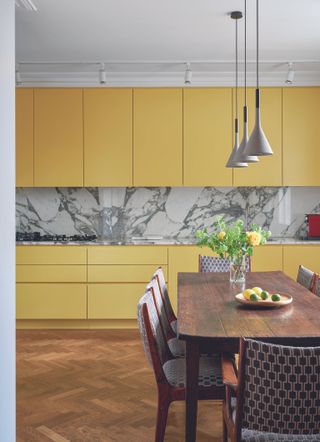 Kitchen with mustard yellow units over grey and white marbled splash back parquet flooring and wooden dining table.